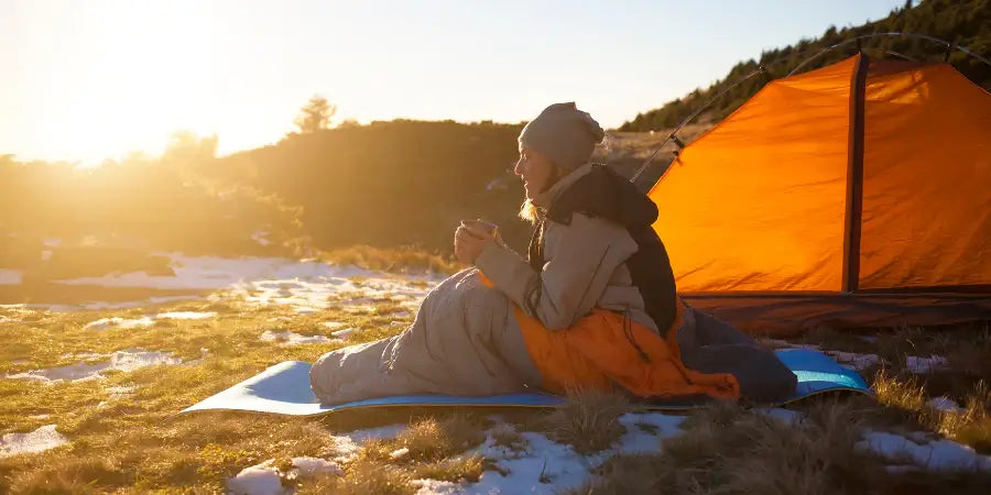 Woman in the wilderness sitting in a warm sleeping bag drinking coffee